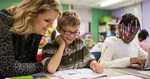 young girl with beads in hair typing at computer sitting next to young boy with glasses writing on paper with teacher leaning over next to him